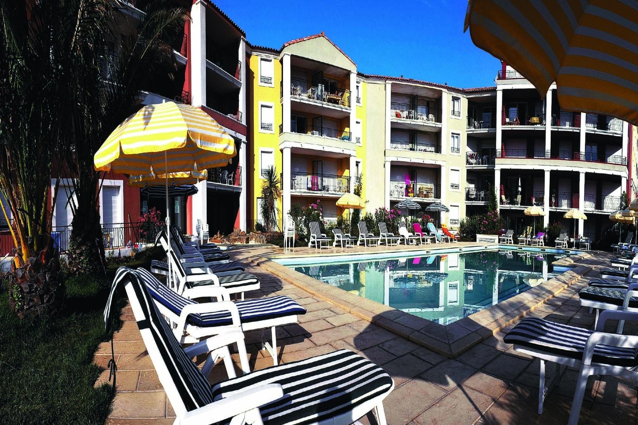 A poolside area with lounge chairs and umbrellas, a pool, and a multi-story building with balconies.