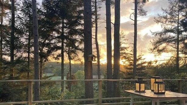 Ulkoterassi, jossa on pöytä ja lyhdyt, ja jota ympäröivät korkeat puut. Terassilta avautuu näkymä auringonlaskuun.