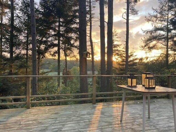 Eine Außenterrasse mit Tisch und Laternen, umgeben von hohen Bäumen und Blick auf den Sonnenuntergang.