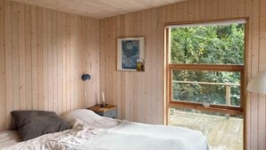 Makuuhuone, jossa on suuri ikkuna, puuseinät, tyynyillä varustettu sänky, yöpöytä ja lamppu.