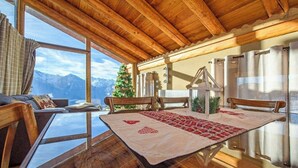 Un intérieur en bois avec une table, des chaises, un sapin de Noël et une vue sur les montagnes, visible à travers de grandes fenêtres.