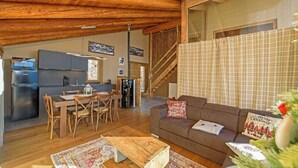 Un salon chaleureux avec un plafond en bois, un canapé agrémenté de coussins aux motifs festifs, un coin repas avec une table et des chaises, et une cuisine équipée d’un réfrigérateur.