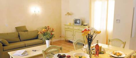 A dining area with a table set for two, a sofa, a TV, and a vase with flowers.