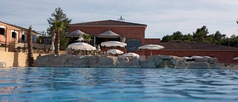 A swimming pool with a building in the background, including outdoor seating and umbrellas.