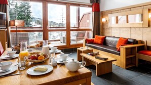 Une pièce chaleureuse avec une table en bois dressée pour le petit-déjeuner, un canapé avec des coussins orange et une grande fenêtre donnant sur des arbres et des montagnes.