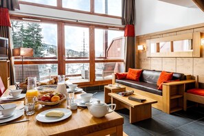 Ein gemütliches Zimmer mit einem Holztisch, gedeckt für das Frühstück, einer Couch mit orangefarbenen Kissen und einem großen Fenster mit Blick auf Bäume und Berge.