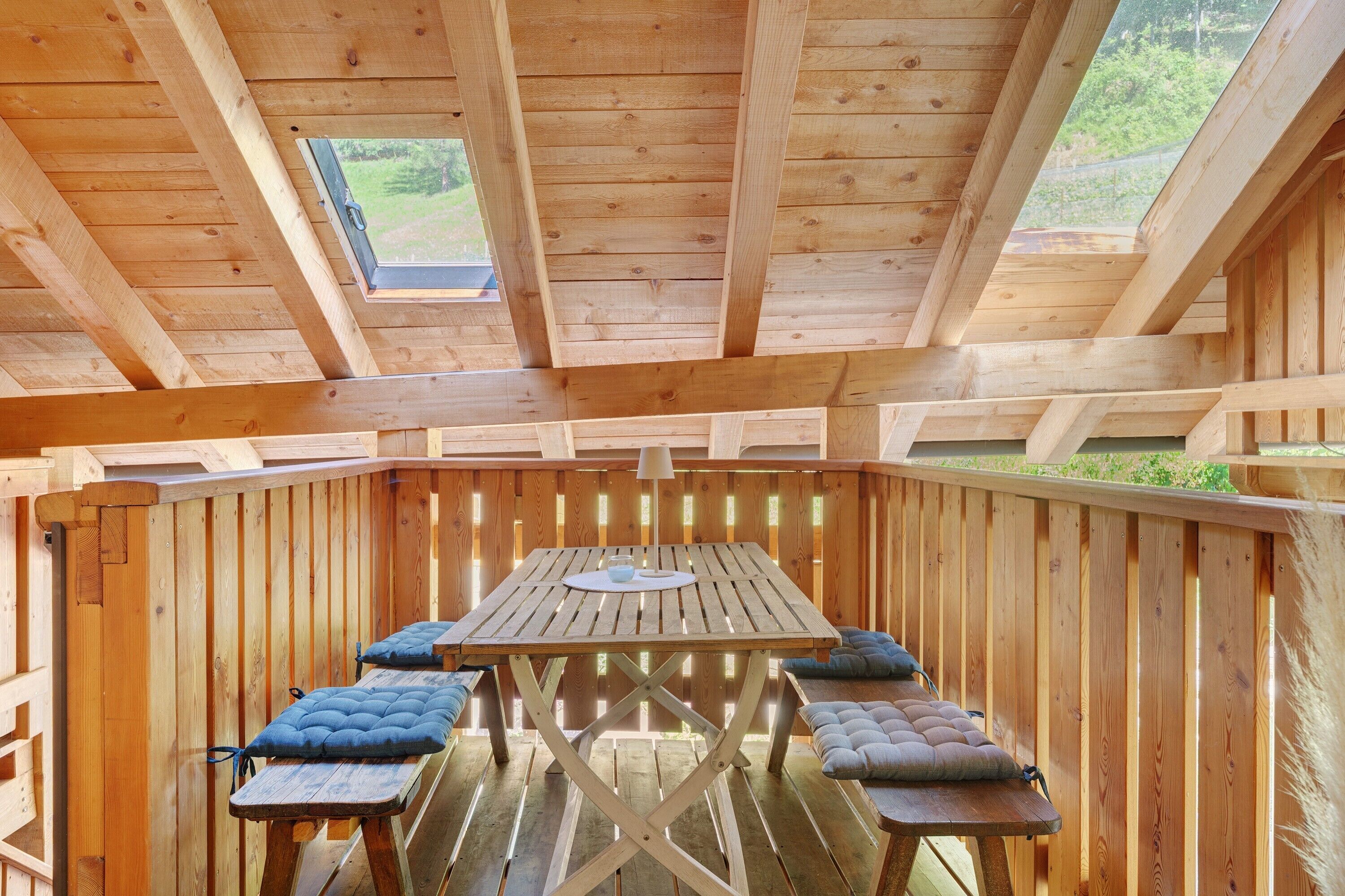 A wooden deck with a table and benches, a skylight, and a view of greenery.