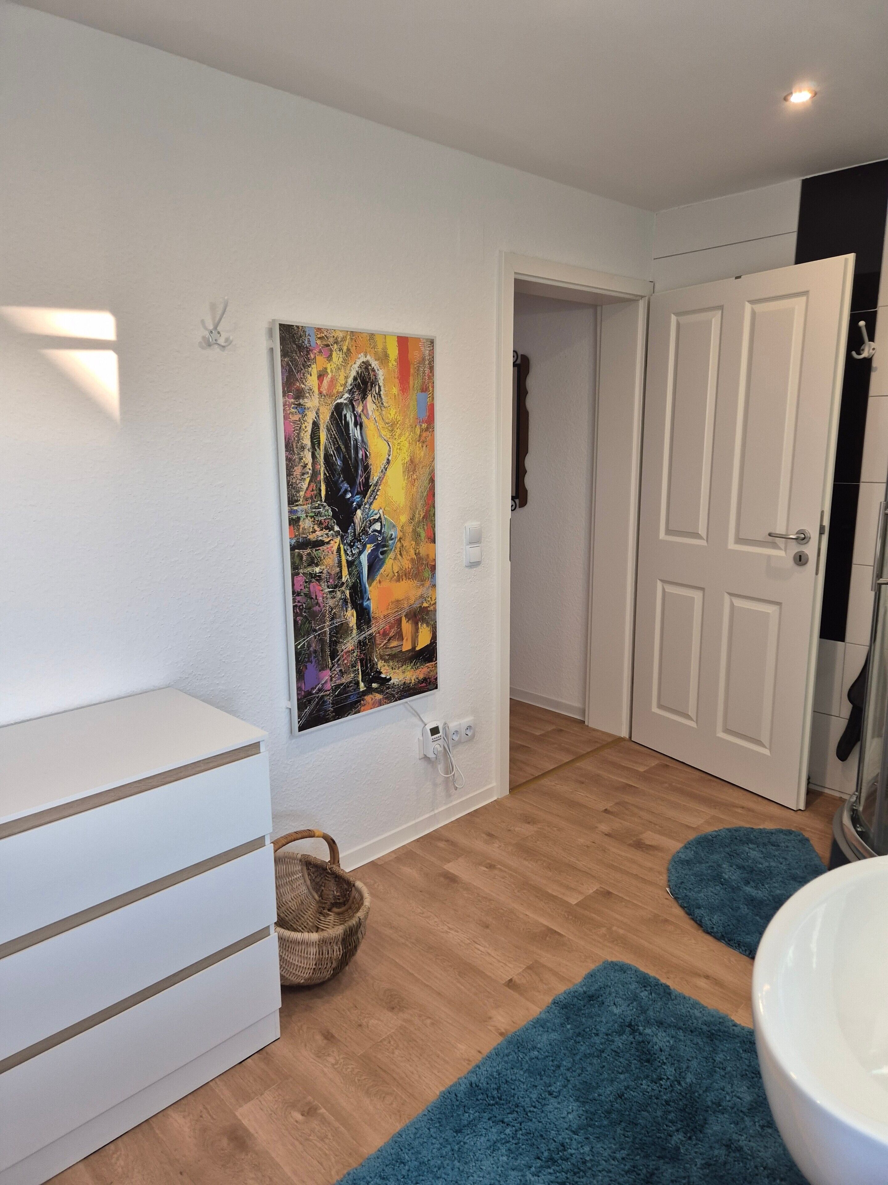 A room with a white dresser, a painting on the wall, a basket, a blue rug, and a white door.