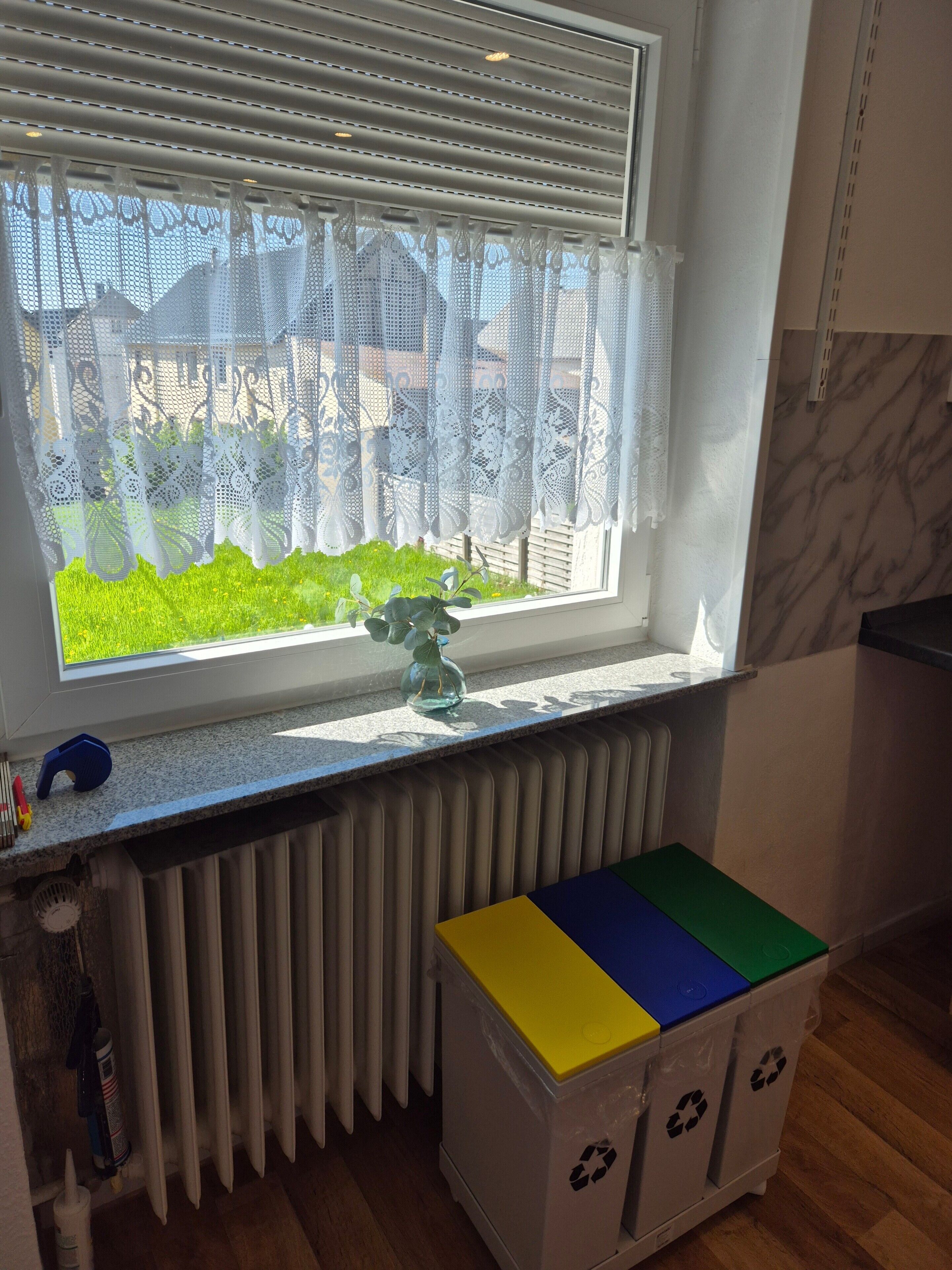 A window with lace curtains, a radiator, a small plant, and a colorful storage box with recycling symbols.