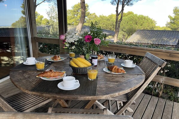 A round wooden table with breakfast items including croissants, bananas, and juice, set on a deck with a view of trees and a house.