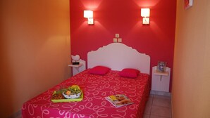 A hotel room with a red wall, a white bed with pink pillows, bedside tables with lamps, and a tray of fruit and a magazine on the bed.