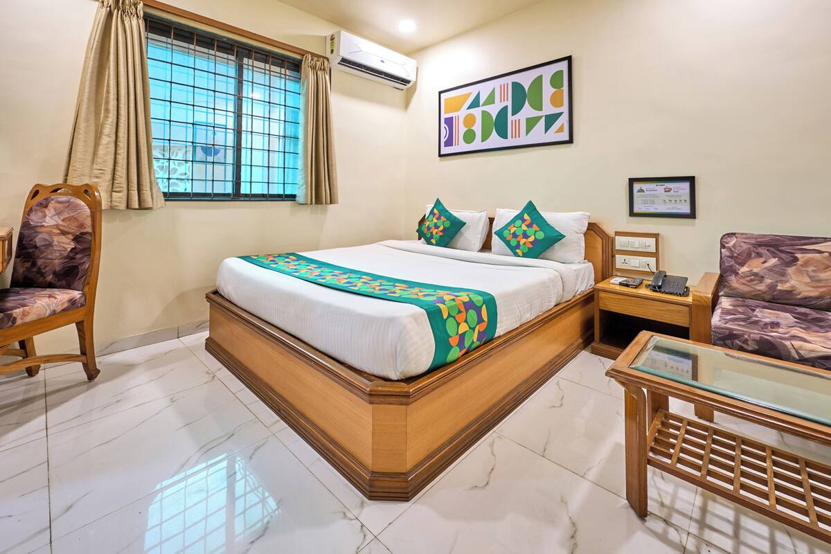 A hotel room with a bed, a chair, a nightstand, a glass table, a window with curtains, and an air conditioner.