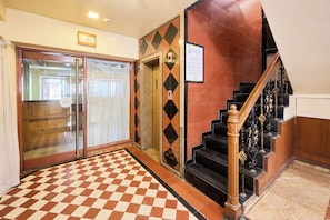 Eine Hotelhalle mit Schachbrettmuster-Boden, einer Treppe mit aufwendiger Geländerarbeit und einem 'PUSH'-Schild an der Tür.