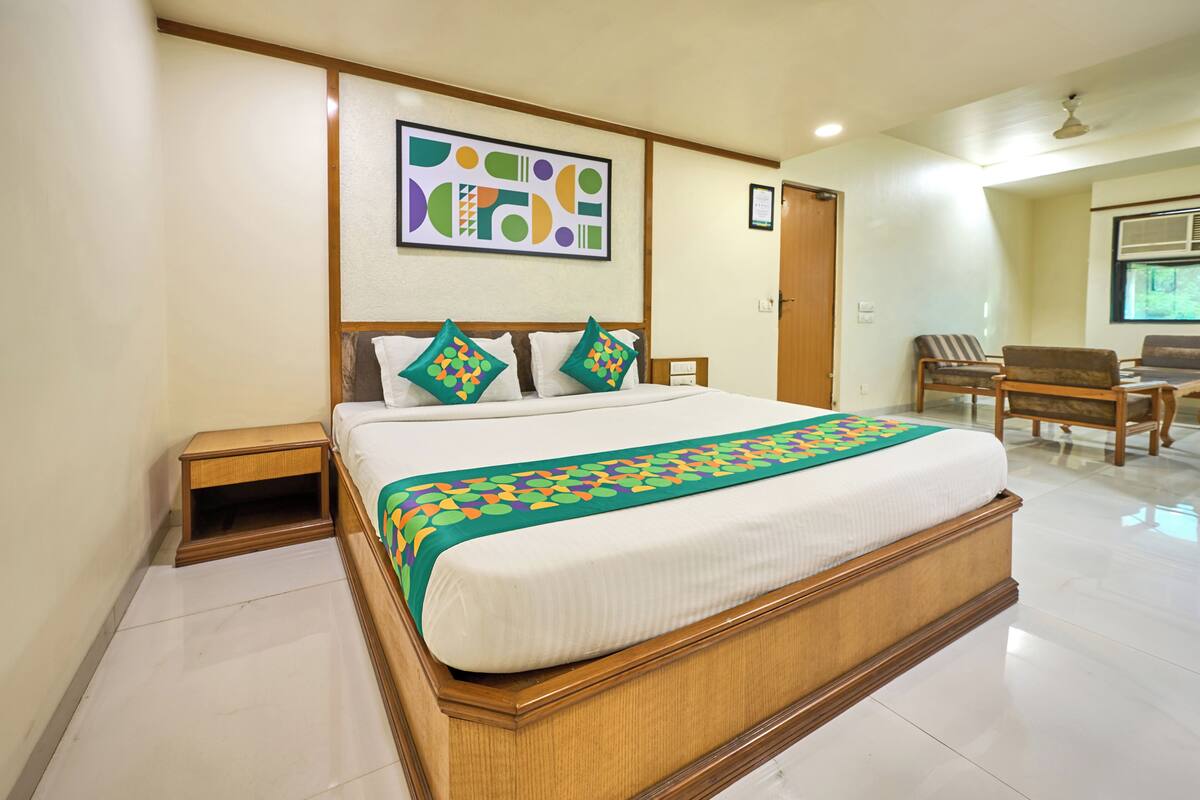 A hotel room with a large bed, a wooden headboard, a small bedside table, a chair, a desk, and a window.