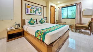 A hotel room with a large bed, a wooden headboard, a bedside table with a kettle, a chair, a window with curtains, and a wall-mounted air conditioner. - Treebo Vice President (Ahmedabad)