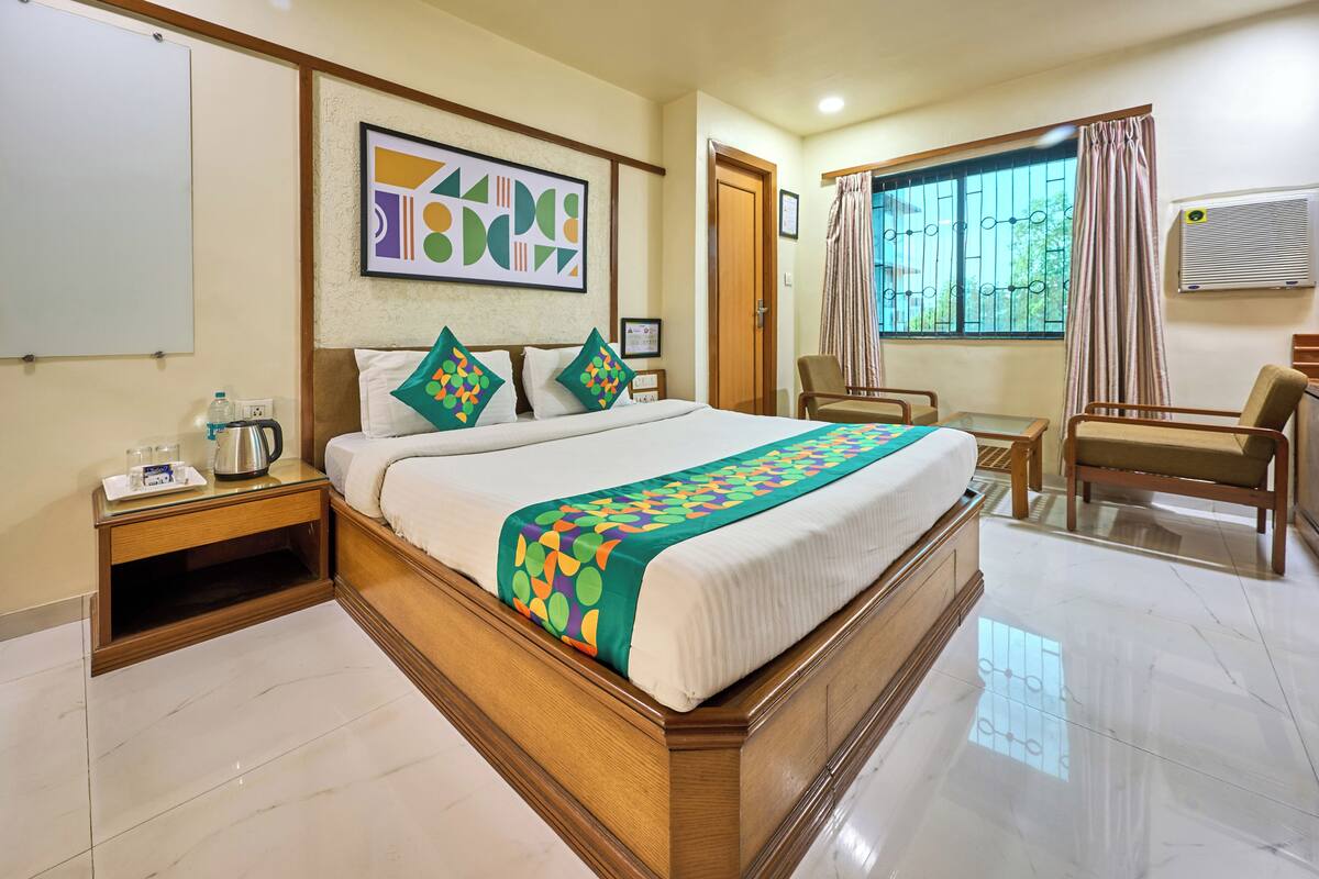 A hotel room with a large bed, a wooden headboard, a bedside table with a kettle, a chair, a window with curtains, and a wall-mounted air conditioner.