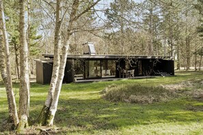 En moderne hytte med glasfacade, omgivet af træer, og med en terrasse med udendørs møbler.