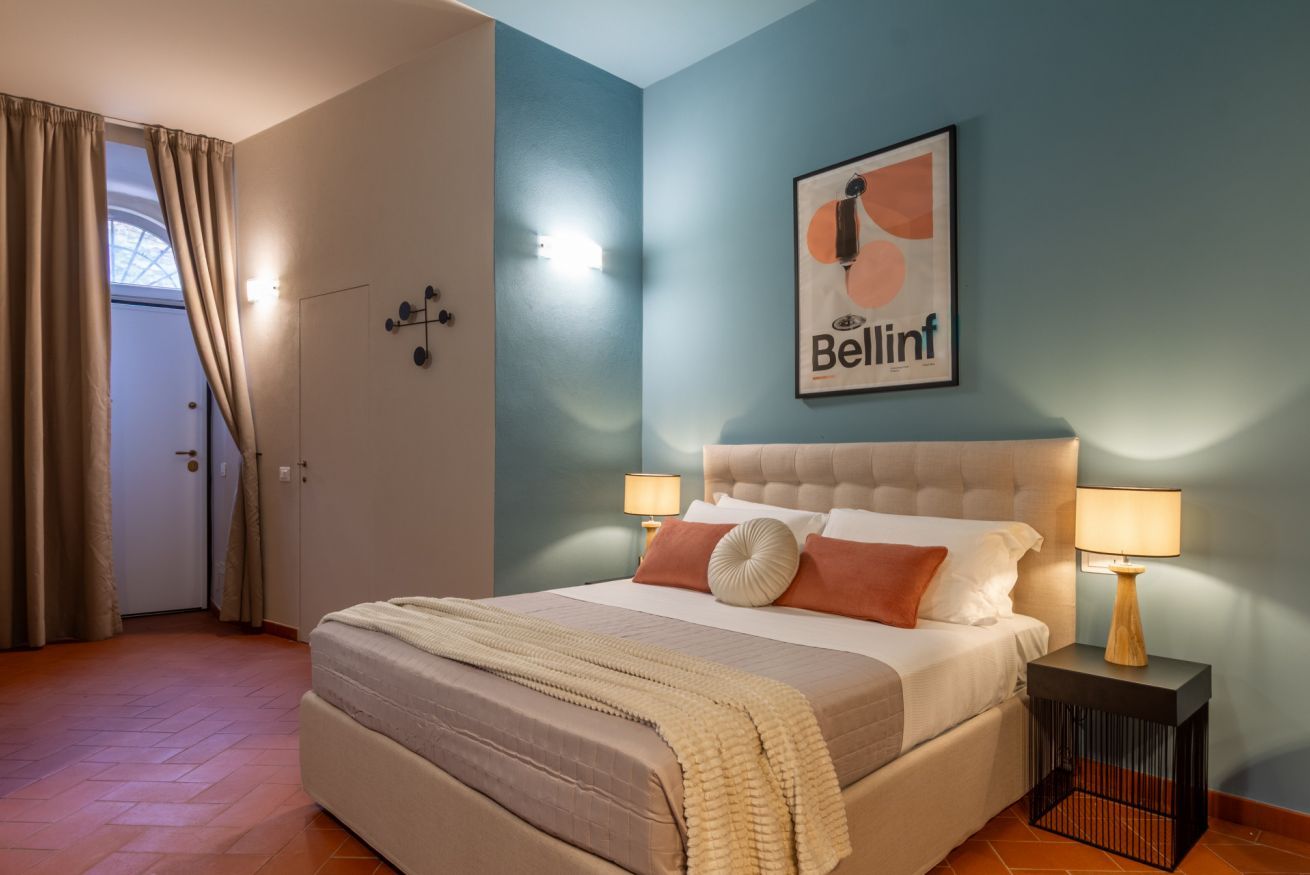 A bedroom with a bed, two bedside lamps, a framed artwork on the wall, and a door with curtains.