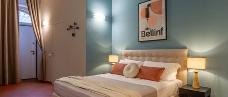 A bedroom with a bed, two bedside lamps, a framed artwork on the wall, and a door with curtains.