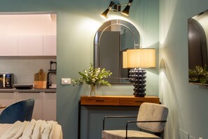 Ein modernes Zimmer mit Bett, Schreibtisch mit Lampe, Spiegel und einer Küchenzeile im Hintergrund.