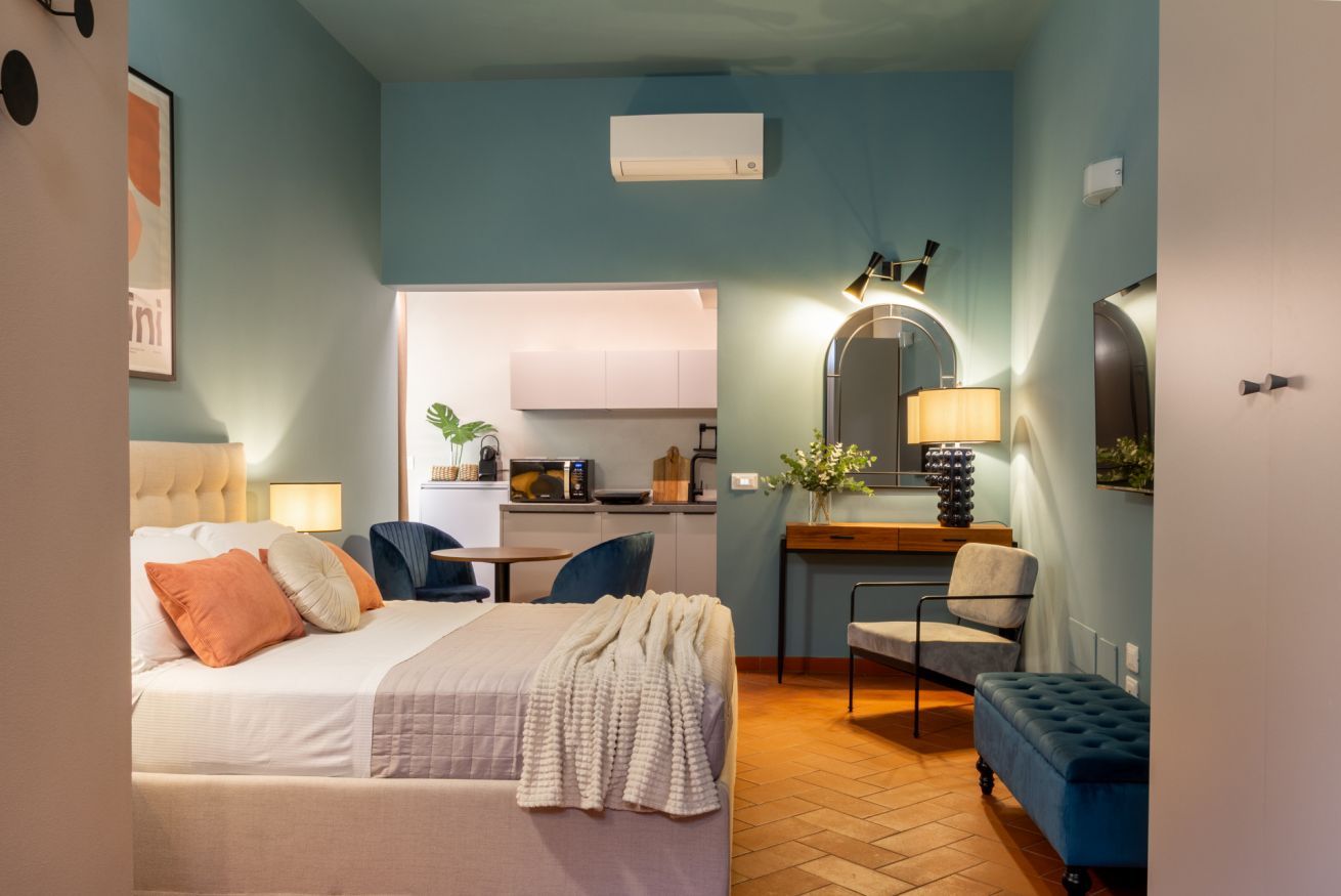 Ein kleines Hotelzimmer mit Bett, Nachttischen, einem Schreibtisch, einem Stuhl und einer kompakten Küchenzeile.