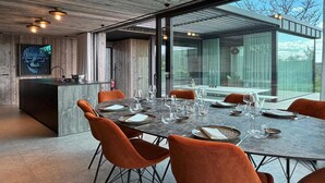 Ein moderner Essbereich mit einem großen Tisch, gedeckt für sechs Personen, umgeben von orangefarbenen Stühlen, sowie eine Küchenzeile mit einer Holz Arbeitsplatte und einem großen Fenster.