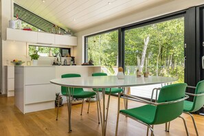 A modern kitchen with a central island, green chairs, and a large window overlooking a forest. - Summer house at Thyholm with fjord view (Thyholm)