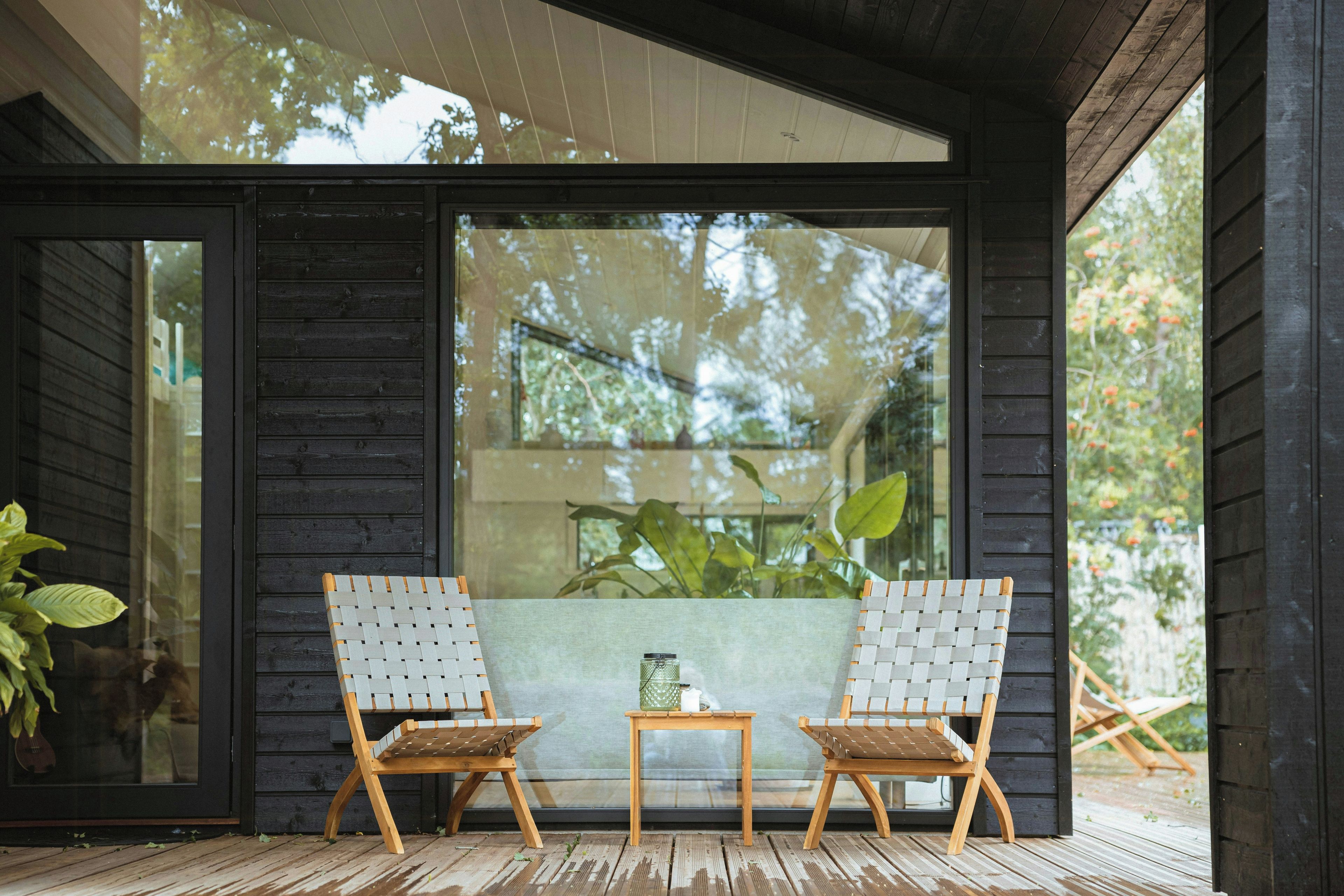 A wooden deck with two chairs and a small table, a glass door leading to a garden, and a view of trees through the door.