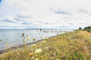 A coastal landscape with a wooden pier extending into calm waters, surrounded by grassy areas and distant land. - Summer house at Thyholm with fjord view (Thyholm)