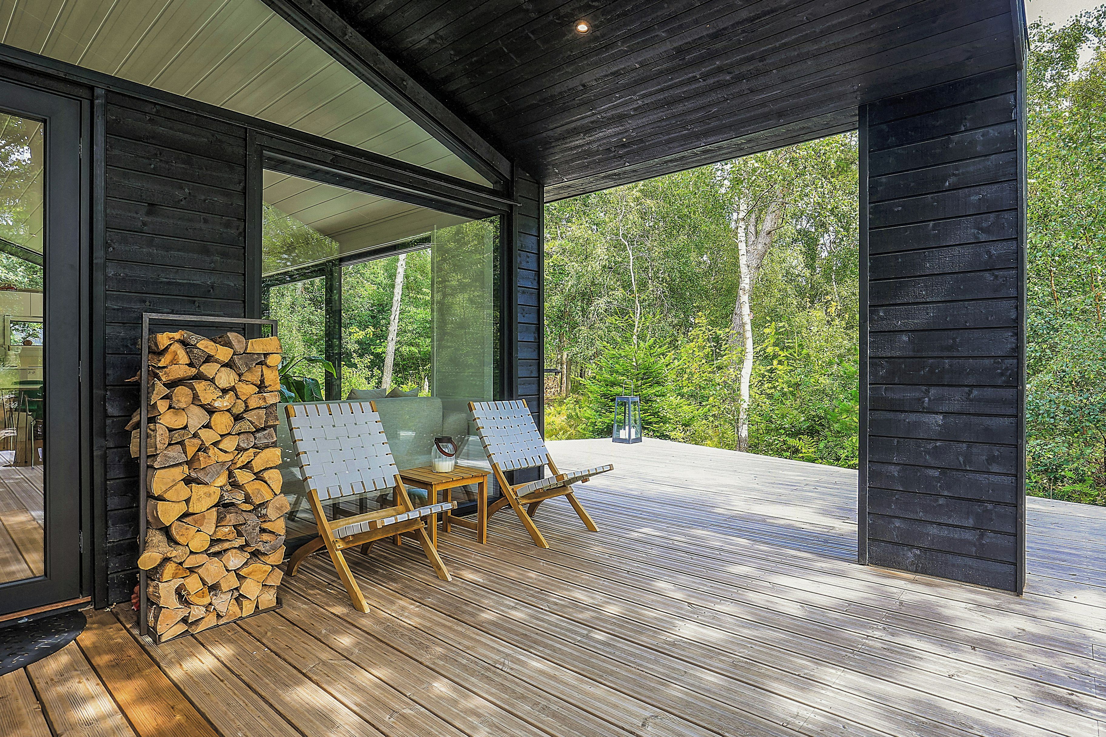 Eine überdachte Holzterrasse mit zwei Loungestühlen, einem Holzscheitestapel und einer Glastür, die zu einem bewaldeten Gebiet führt.