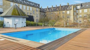 Una piscina amb un tram de fusta, un cobert petit i un edifici de diversos pisos al fons.