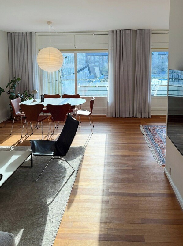 Un salon moderne avec une table à manger ronde, des chaises rouges, un canapé noir, une grande fenêtre avec des rideaux et un luminaire suspendu.