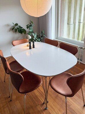 Une table ronde entourée de six chaises marron, un luminaire suspendu et une plante en pot, le tout dans une pièce au plancher en bois et dotée d’une fenêtre.