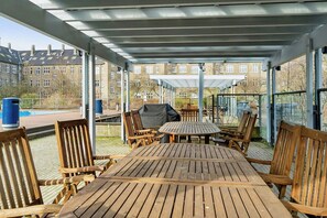 Une terrasse extérieure couverte, aménagée avec du mobilier en bois, un barbecue et offrant une vue sur des immeubles d’habitation.