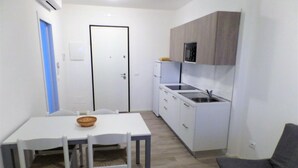 Una cocina compacta con armarios blancos, microondas integrado y un lavamanos. Hay una mesa de comedor con sillas y un sofá gris en la sala de estar.