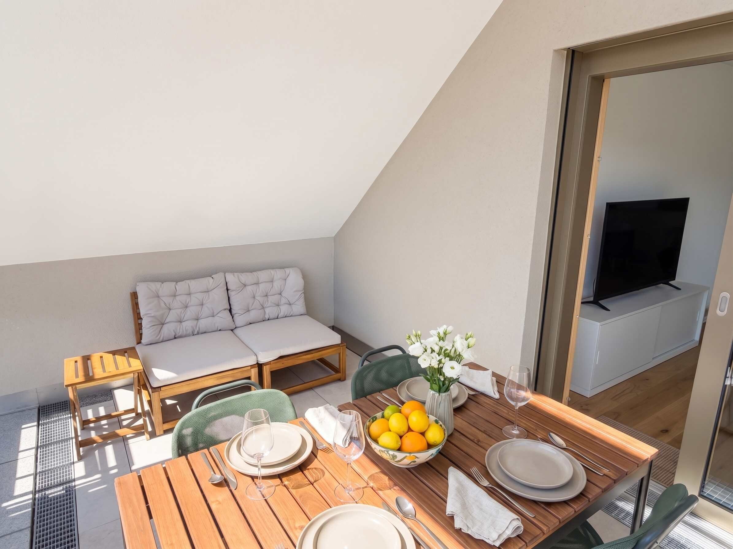 A dining area with a wooden table set for two, a sofa, and a TV in the background.