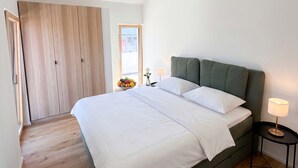 A bedroom with a bed, a nightstand, a lamp, a window, and a wooden wardrobe.