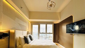 A modern bedroom with a large bed, a TV mounted on the wall, and a city view through the window. - Cozy Sunlit Lounge w/ Balcony City View Uptown BGC (Taguig)