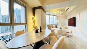 A modern hotel room with a large round table, comfortable seating, a sofa, a TV, and large windows offering a city view. - Cozy Sunlit Lounge w/ Balcony City View Uptown BGC (Taguig)
