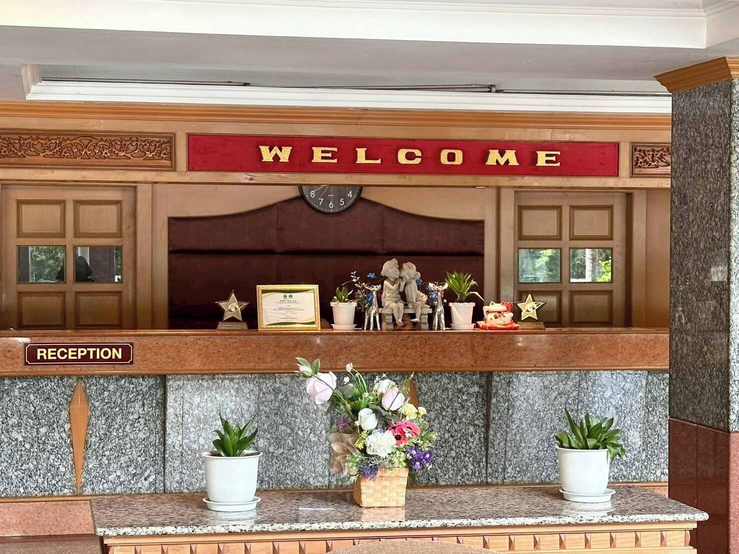 A reception desk with a 'WELCOME' sign, a clock, framed certificates, and potted plants.
