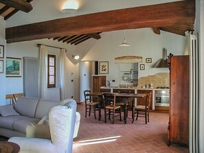 A living room with a sofa, dining table, and chairs, a kitchen with a stove and sink, and wooden beams on the ceiling. - Stone house apartment with pool in Tuscan hills (Asciano)