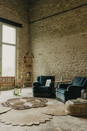 A rustic room with a stone wall, a ladder with lights, two dark blue armchairs, a woven rug, and a small round table with a book. - Charming 1920s house in Picardy, 2h from Paris (Sainte-Croix)