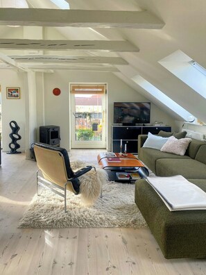 A loft-style living room with a high ceiling, a sofa, a chair, a TV, and a view through a large window. - Penthouse with private garden (København)