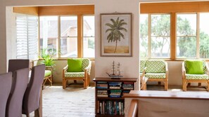 A room with a bookshelf, two green cushioned chairs, a wooden table, and a framed picture of a palm tree. - Sandilands 1 - Lifestyle Yamba (YAMBA)