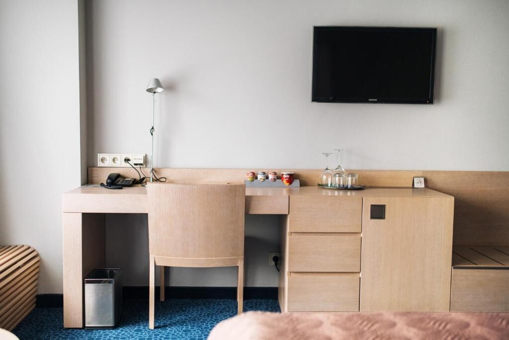 Et hotelværelse med et træskrivebord, en stol, et tv ophængt på væggen, en lampe, en skraldespand og en lille hylde med genstande.