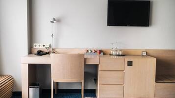 Et hotelværelse med et træskrivebord, en stol, et tv ophængt på væggen, en lampe, en skraldespand og en lille hylde med genstande.
