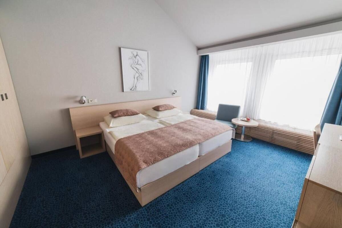 Una habitación de hotel con cama, mesitas de noche, una pequeña mesa redonda, una silla, un ventanal con cortinas y una obra de arte colgada en la pared.
