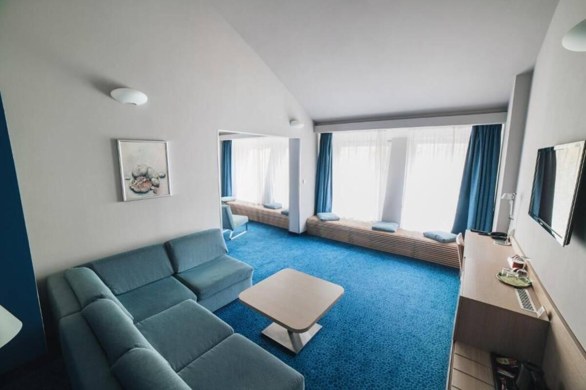Una habitación de hotel moderna con alfombra azul, un sofá, una mesita, televisión y ventana con cortinas.