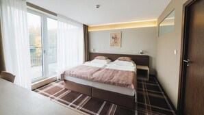 A modern hotel room with a large bed, a bedside table, a chair, a view through a sliding door, and a small artwork on the wall.