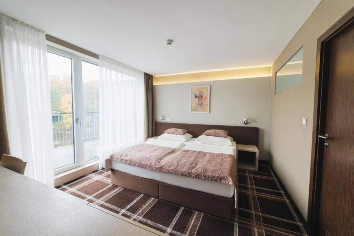 Una habitación de hotel moderna con una cama grande, una mesita de noche, una silla, vistas a través de una puerta corredera y una pequeña obra de arte en la pared.